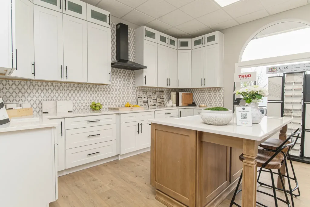 A bright kitchen showroom with white cabinetry, a wooden island, and decorative backsplash tiles showcasing cabinet hardware and quartz countertop samples