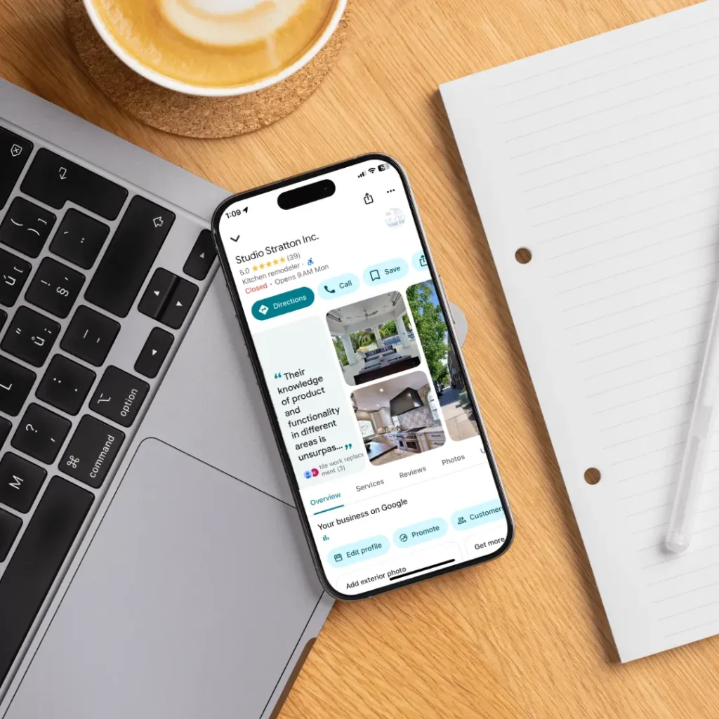 Mobile phone displaying Studio Stratton's Google Business Profile with reviews and images of kitchen and bath designs, placed next to a laptop and coffee cup on a wooden desk